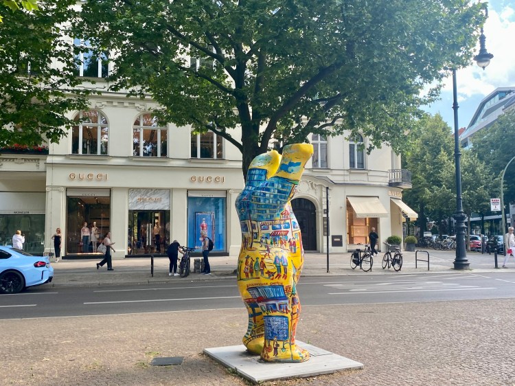 Bunte Bärenstatue auf einer Straße vor Luxusgeschäften, umgeben von Bäumen und Passanten.
