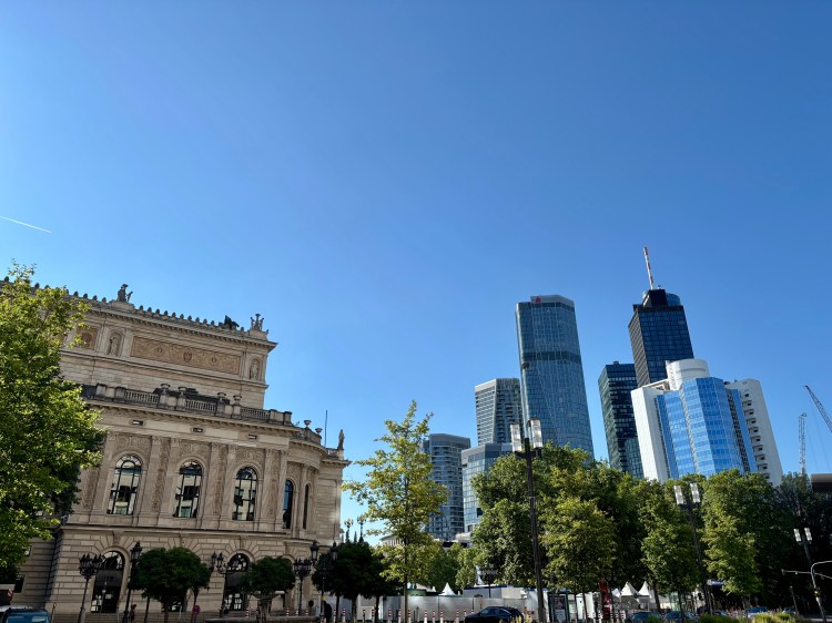 Ein historisches Gebäude im Vordergrund mit einer eleganten Fassade, umgeben von Bäumen, während im Hintergrund moderne Wolkenkratzer unter einem strahlend blauen Himmel stehen.