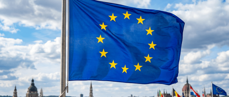 European Union flag with twelve yellow stars on blue background flying above urban buildings