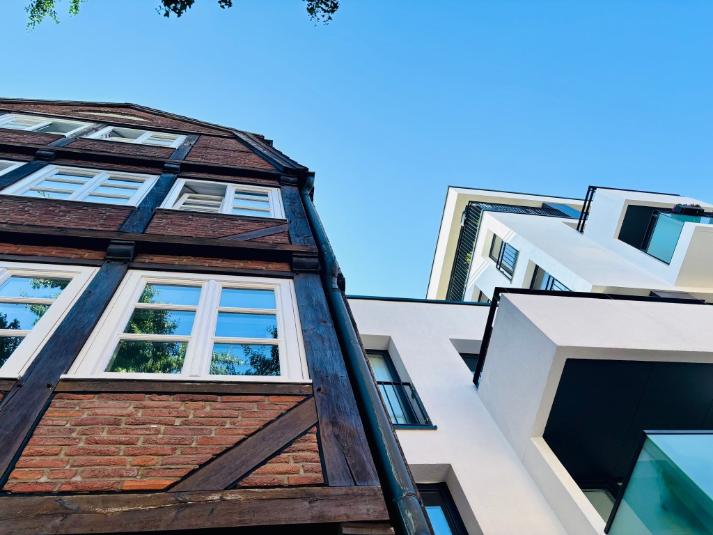 Aufnahme von zwei Gebäuden aus der Froschperspektive, eines ist traditionell mit roten Ziegeln und Fachwerk, das andere modern und weiß mit Balkonen, vor blauem Himmel.